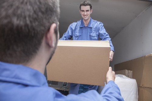 A professional removal worker in a blue uniform is smiling as he hands a large cardboard box to a colleague inside the back of a removals van, which is filled with various packed boxes and wrapped belongings. The interior of the van shows shelves and a plain white ceiling, suggesting a residential move nearby in Kennington or the surrounding London areas. The worker's face is visible with a friendly expression, and the focus is on the transfer of the box, highlighting the careful handling involved in house removals. The scene captures the process of loading household items such as cardboard boxes, which vary in size, onto the vehicle, emphasizing efficient packing and transportation. Natural lighting illuminates the scene, reflecting a typical day of house removals, with the environment indicating a suburban street or driveway setup. The attention to the professional service offered by Kennington Man with Van is subtly supported by this image, illustrating a reliable and organized moving process in South London postcode areas.