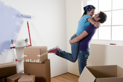 A young couple in a bright, minimally furnished room are engaged in moving house, with the man lifting the woman off the floor while she wraps her arms around his shoulders. The woman is wearing a blue top and jeans, and the man is dressed in a purple t-shirt and jeans. They appear happy and are smiling during the moment. The room features a large window allowing natural light to illuminate the scene, with light-colored walls and wooden flooring. In the background, there are stacked cardboard moving boxes of various sizes, some closed and taped, and one open box on the floor. Near the corner, there is a white furniture cover or protective sheet, partially rolled up, hanging on the wall. The overall environment suggests a house in the process of relocation, with household belongings prepared for moving, supporting professional house removal services such as those offered by Kennington Man with Van in the local area of Kennington or nearby postcode. The scene captures an active moment during a house move, emphasizing the household items, their textures, and the residential setting involved in local removals.