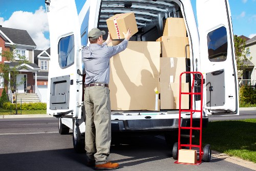 A man in casual work attire, including a grey shirt and beige trousers, is seen loading cardboard moving boxes into the back of a white removals van parked on a residential street. The van's rear doors are open, revealing a large, empty cargo space with some boxes already inside. Nearby, a red hand truck with two wheels is positioned on the pavement, holding a small box at its base. In the background, a suburban neighbourhood with modern houses, manicured lawns, and a clear blue sky is visible, indicating a daytime move in a quiet residential area within the vicinity of Kennington or nearby London postcode areas. The scene captures an organized house removal process, with attention to packing and transportation of household belongings, consistent with professional removals services such as those offered by Kennington Man with Van.