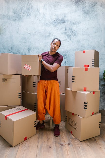 A professional removal service in progress inside a bright, minimally furnished room with three large arched windows allowing natural daylight. A mover, dressed in dark uniform, is carrying two medium