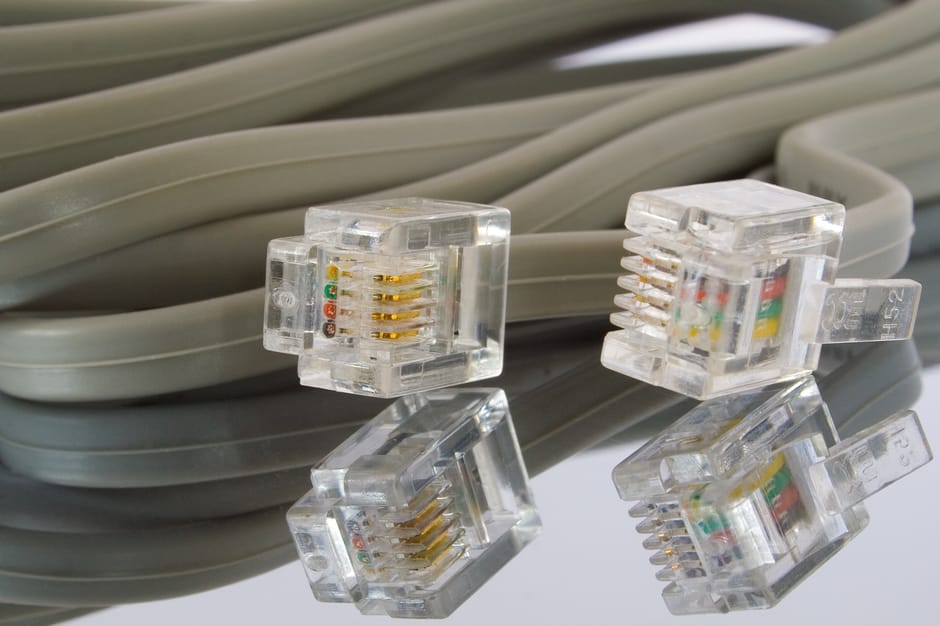 Close-up of a coiled gray Ethernet cable with four transparent RJ45 connectors placed on top, showing gold-plated pins and multicoloured wire arrangements inside. The cable is situated on a reflective