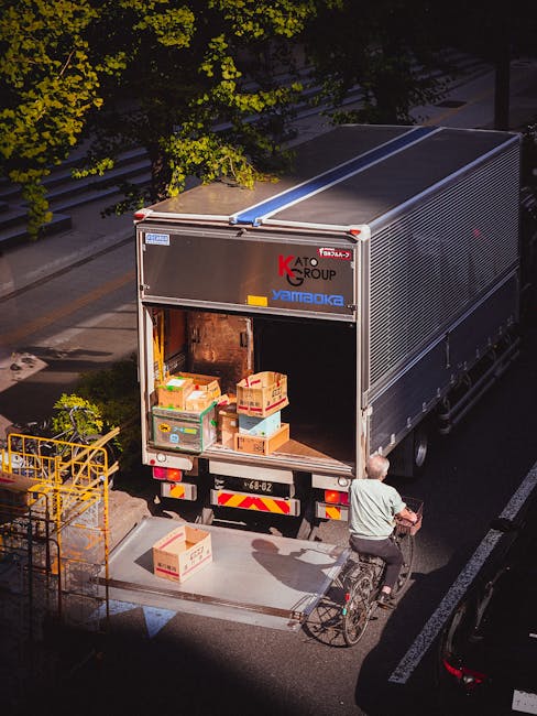 A man wearing a black face mask and a blue long-sleeve shirt is standing outside the open rear doors of a white moving van, holding three cardboard boxes of varying sizes, with the largest at the bott