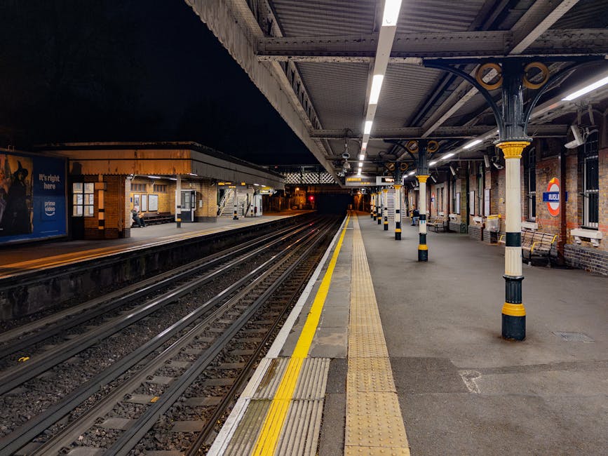 A nighttime view of Kennington station platform, featuring train tracks running parallel to the platform edge, which is marked by a yellow safety line. The platform includes a textured tactile paving
