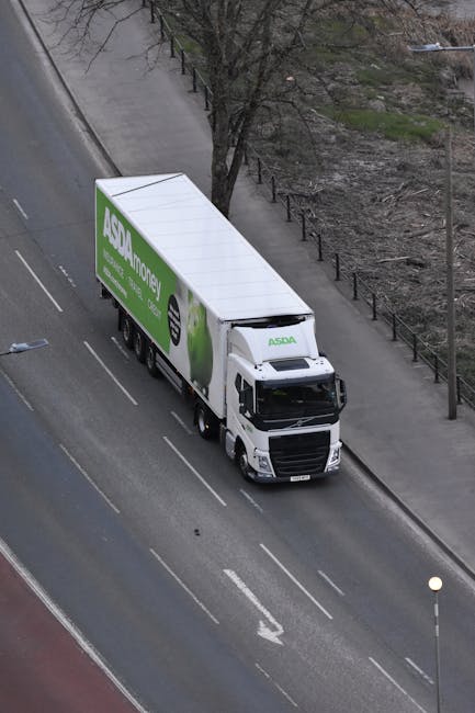 An aerial view of a white moving truck with an ASDA Money advertisement on its side, parked on a paved street adjacent to a sidewalk and a row of metal railings. The truck is positioned parallel to th